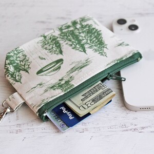 Puede incluir: Una peque&ntilde;a bolsa verde con cremallera, con fondo blanco y un patr&oacute;n verde y blanco de &aacute;rboles y un platillo volante. La bolsa tiene un llavero plateado y est&aacute; abierta para revelar una tarjeta de cr&eacute;dito y dinero en efectivo en su interior.