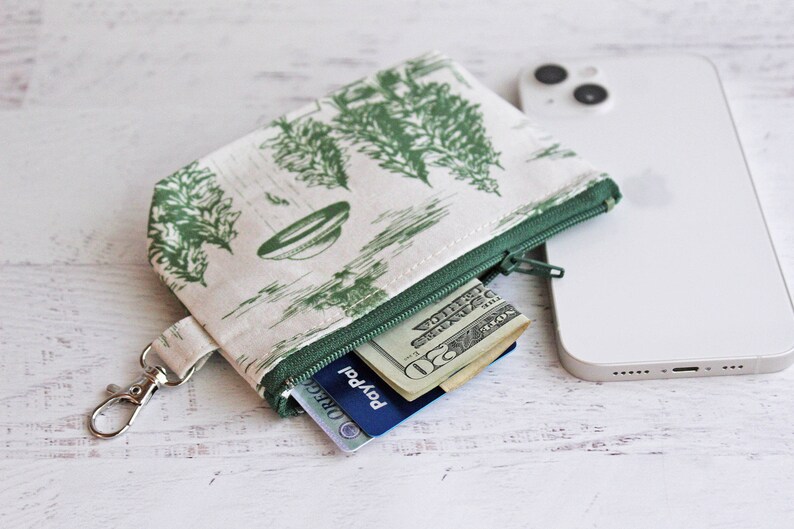 Puede incluir: Una peque&ntilde;a bolsa verde con cremallera, con fondo blanco y un patr&oacute;n verde y blanco de &aacute;rboles y un platillo volante. La bolsa tiene un llavero plateado y est&aacute; abierta para revelar una tarjeta de cr&eacute;dito y dinero en efectivo en su interior.
