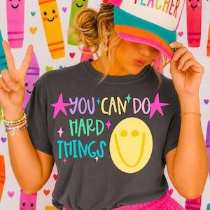 May include: A charcoal gray t-shirt featuring the text YOU CAN DO HARD THINGS in colorful letters with a yellow smiley face and pink stars. The shirt is paired with a white and teal trucker hat with the word TEACHER and bright pink shorts.