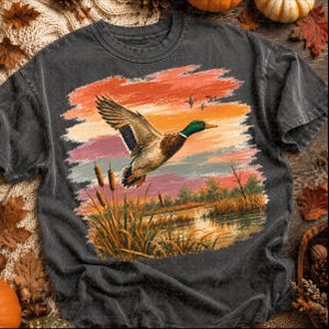 May include: A charcoal gray t-shirt featuring a graphic print of a mallard duck in flight over a marshy landscape. The scene includes tall cattails in the foreground, a calm body of water, and a vibrant sunset sky with orange and purple hues.