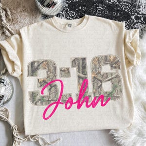 Könnte beinhalten: Ein cremefarbenes Kurzarm-T-Shirt mit dem Text 3:16 in einem Tarnmuster, wobei das Wort John in einer leuchtend pinken Schreibschrift über die Zahlen gelegt ist. Das T-Shirt hat einen Rundhalsausschnitt und ein lockeres Design.
