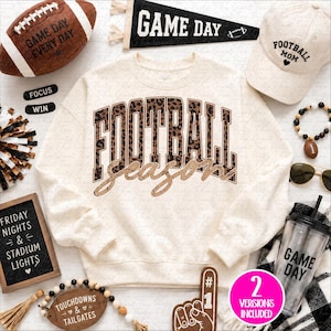 Puede incluir: Una sudadera color crema con la palabra FOOTBALL en estampado de leopardo marrón y la palabra season en letra dorada brillante. La imagen incluye un balón de fútbol marrón, un banderín negro, una gorra beige, una pulsera de cuentas y un vaso.
