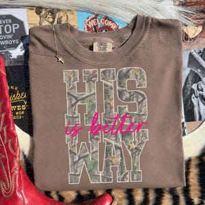 K&ouml;nnte beinhalten: Ein braunes Kurzarm-T-Shirt mit dem Text HIS WAY in gro&szlig;en Blockbuchstaben, die mit einem Tarnmuster gef&uuml;llt sind. Die W&ouml;rter is better sind in einer leuchtend pinken Schreibschrift &uuml;ber die Mitte des Tarntextes gedruckt.