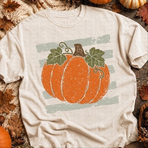 May include: A cream-colored short-sleeve t-shirt featuring a large, distressed-style orange pumpkin graphic with green leaves and vines, centered over a background of horizontal, faded sage green brushstroke stripes.