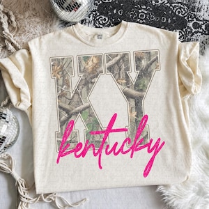 May include: A cream-colored short-sleeve t-shirt featuring the letters KY in a block font filled with a forest camouflage pattern. The word Kentucky is printed across the bottom of the letters in a bright pink, cursive, glitter-style font.