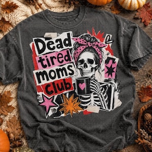 May include: A charcoal gray t-shirt featuring a graphic of a skeleton wearing a pink polka-dot headband and holding a black mug with a pink heart. The text reads Dead tired moms club, surrounded by pink and orange star shapes and paper scraps.