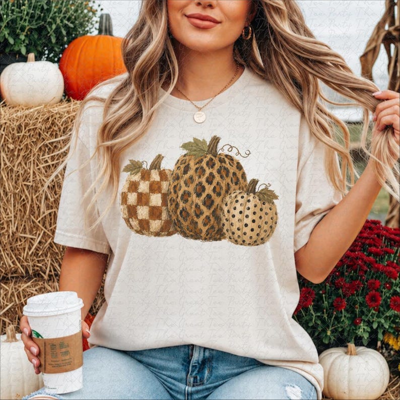 May include: A beige short-sleeve t-shirt featuring a graphic print of three pumpkins. The left pumpkin has a tan and white checkered pattern, the center pumpkin has a brown leopard print, and the right pumpkin has a tan and black polka dot pattern.