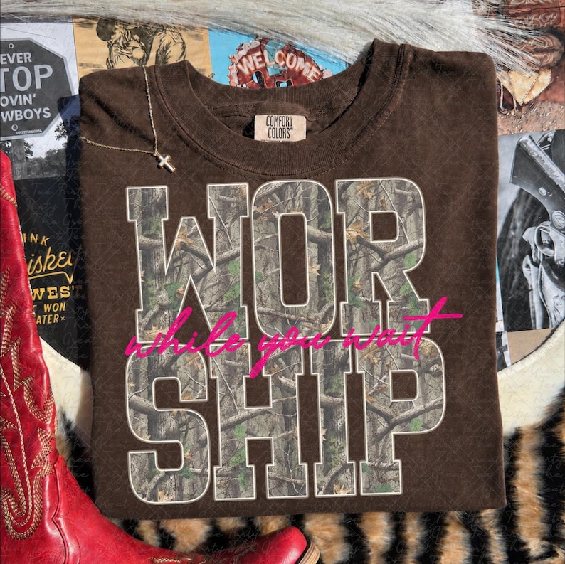 May include: A brown short-sleeved t-shirt with a Comfort Colors label at the neck. The shirt features the word WORSHIP in large block letters filled with a camouflage pattern, overlaid with the pink cursive text while you wait.
