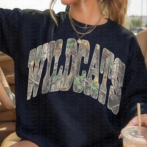 May include: A black long-sleeve sweatshirt featuring the word WILDCATS in a large, arched, collegiate-style font. The letters are filled with a realistic forest camouflage pattern consisting of green leaves, brown branches, and tree bark textures.