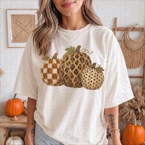 May include: An off-white short-sleeve t-shirt featuring a graphic print of three pumpkins. The left pumpkin has a brown and white checkered pattern, the center pumpkin has a brown leopard print, and the right pumpkin has a brown polka dot pattern.