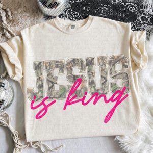 May include: A cream-colored short-sleeve t-shirt featuring the text JESUS in a block font with a camouflage pattern fill, layered with the word is king in a bright pink cursive script font across the center of the garment.