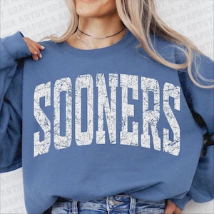 May include: A blue sweatshirt with the word "SOONERS" in distressed white lettering. The sweatshirt is a solid blue color and has a crew neck. The text is in a bold, collegiate-style font.