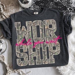 May include: A charcoal gray short-sleeve t-shirt featuring the word WORSHIP in large block letters filled with a camouflage tree branch pattern. The phrase while you wait is printed across the center in a bright pink cursive script font.