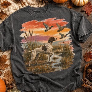 May include: A charcoal gray short-sleeve t-shirt featuring a graphic print of a brown and white spotted hunting dog standing in a marsh with tall reeds. Three ducks are flying in the sky above against a sunset background of orange and pink.