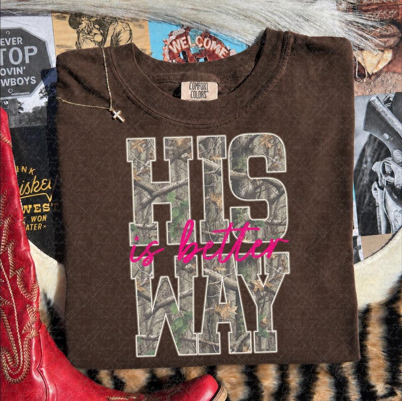 K&ouml;nnte beinhalten: Ein braunes Kurzarm-T-Shirt mit Rundhalsausschnitt und einem Comfort Colors Markenetikett. Auf der Vorderseite befindet sich der Text HIS WAY in gro&szlig;en Blockbuchstaben mit Camouflage-Muster sowie die Worte is better in pinker Schreibschrift.