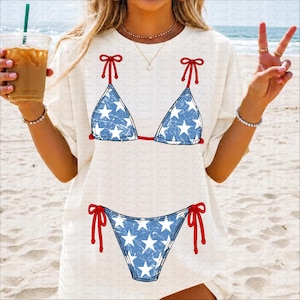 May include: White t-shirt featuring a blue and white star-patterned bikini design with red bow accents. The bikini top and bottom are printed on the shirt, creating a patriotic summer look. The shirt is worn on a beach.