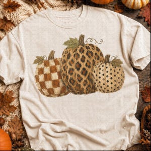 May include: A cream-colored short-sleeve t-shirt featuring a graphic of three pumpkins. The left pumpkin has a brown and cream checkerboard pattern, the center pumpkin has a brown leopard print, and the right pumpkin has a cream and black polka dot pattern.
