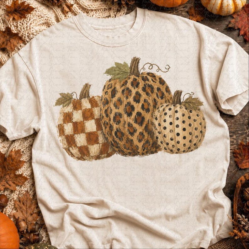 May include: A cream-colored short-sleeve t-shirt featuring a graphic of three pumpkins. The left pumpkin has a brown and cream checkerboard pattern, the center pumpkin has a brown leopard print, and the right pumpkin has a cream and black polka dot pattern.