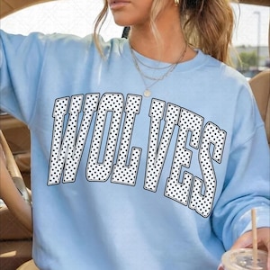May include: A light blue crewneck sweatshirt featuring the word WOLVES printed in a large, arched collegiate font. The letters are white with a black polka dot pattern and a thin black outline, creating a bold graphic design on the chest.