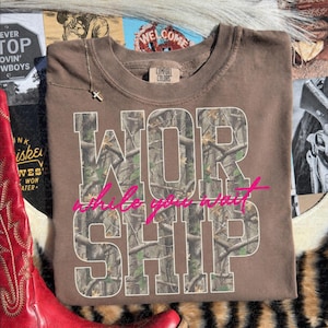 May include: A brown short-sleeved t-shirt with a crew neckline and a Comfort Colors label. The front features the word WORSHIP in large block letters filled with a camouflage tree pattern, overlaid with the pink cursive text while you wait.