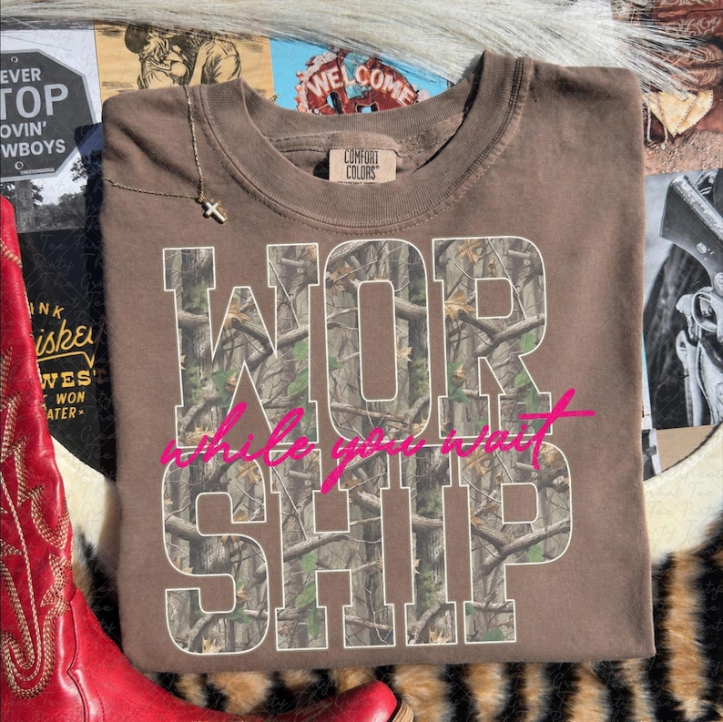 May include: A brown short-sleeved t-shirt with a crew neckline and a Comfort Colors label. The front features the word WORSHIP in large block letters filled with a camouflage tree pattern, overlaid with the pink cursive text while you wait.