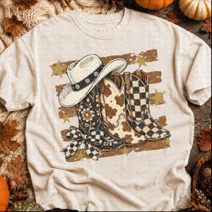 Könnte beinhalten: Ein cremefarbenes T-Shirt mit einer Grafik, die einen weißen Cowboyhut auf drei Cowboystiefeln zeigt. Die Stiefel haben Blumenmuster, Kuhflecken und schwarz-weiße Karomuster. Eine karierte Schleife ist am vorderen Stiefel befestigt.
