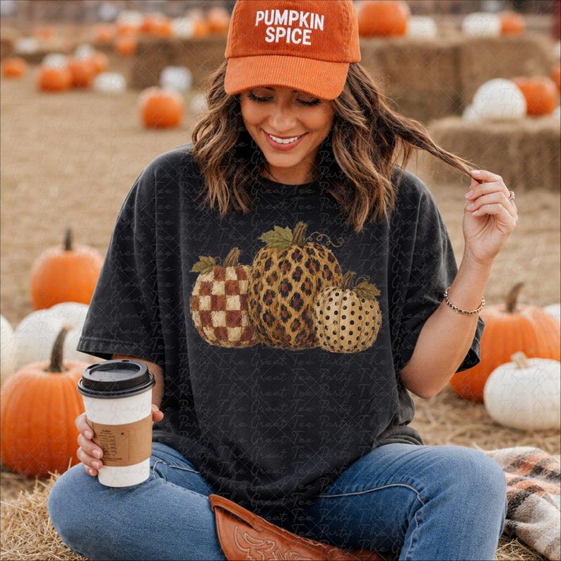 May include: A charcoal gray t-shirt featuring a graphic of three pumpkins with different patterns: a brown checkered pumpkin, a leopard print pumpkin, and a tan polka dot pumpkin. The shirt is paired with an orange baseball cap with PUMPKIN SPICE text.