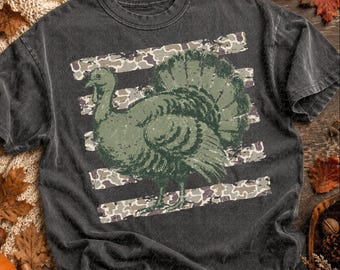 May include: A charcoal gray short-sleeve t-shirt featuring a distressed olive green graphic of a turkey standing in front of horizontal stripes with a camouflage pattern. The shirt has a crew neckline and a faded, vintage-inspired fabric texture.