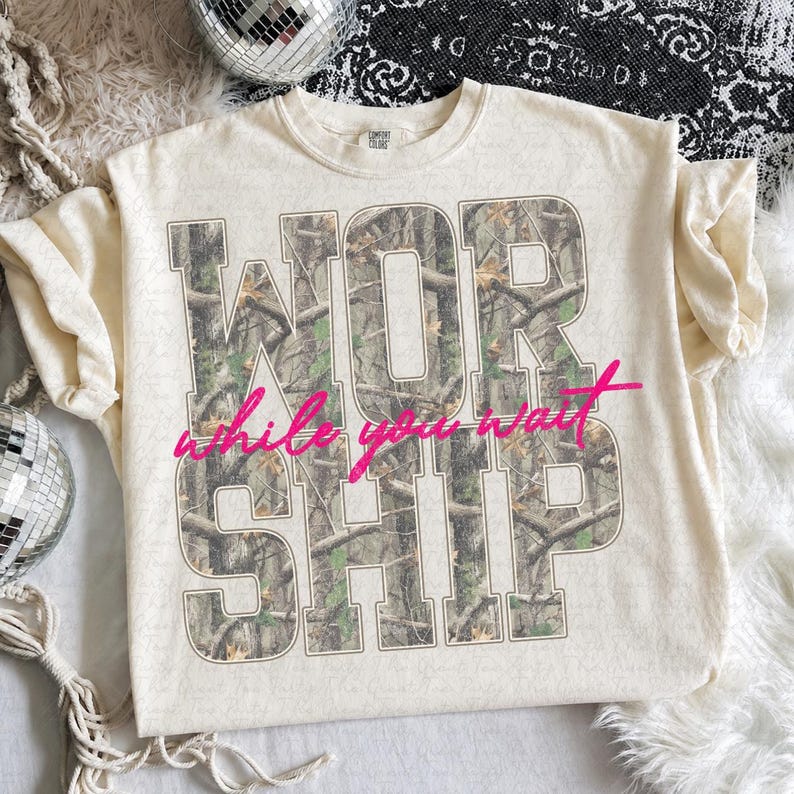 May include: A cream-colored short-sleeve t-shirt featuring the word WORSHIP in large block letters filled with a green and brown forest camouflage pattern. The phrase while you wait is printed across the center in bright pink cursive script.