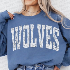 May include: A blue sweatshirt with the word "WOLVES" printed in large, distressed white letters. The sweatshirt is a solid blue color and has a crew neck. The image shows the upper body of a person wearing the sweatshirt.