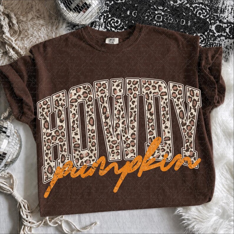 Puede incluir: Una camiseta de manga corta de color marr&oacute;n oscuro con la palabra HOWDY en letras grandes de bloque con un estampado de leopardo beige y negro. Debajo de la palabra HOWDY, la palabra pumpkin est&aacute; impresa en una fuente cursiva naranja desgastada.