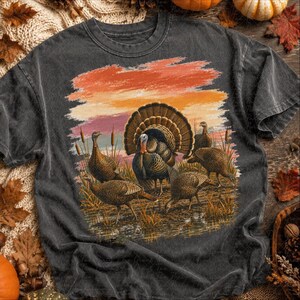 May include: A charcoal gray short-sleeve t-shirt featuring a graphic print of five turkeys in a marshy field. The scene includes tall grasses and a sunset sky with orange, pink, and yellow hues. The turkeys are depicted in shades of brown and black.