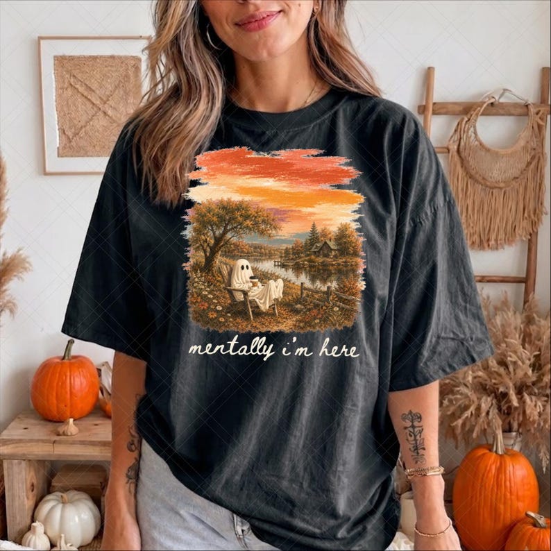 May include: A black short-sleeve t-shirt featuring a graphic print of a ghost sitting in a chair by a lake during sunset. The text "mentally i'm here" is printed in white cursive script below the illustration, which includes trees and flowers.