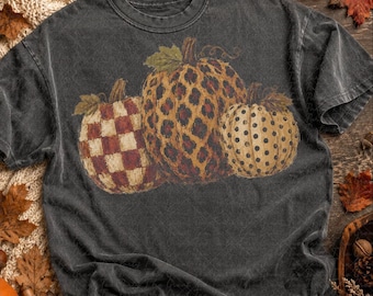 Puede incluir: Una camiseta de manga corta gris carbón con un estampado gráfico de tres calabazas. La calabaza de la izquierda tiene un patrón de cuadros rojos y blancos, la central un estampado de leopardo marrón y la derecha un diseño de lunares.