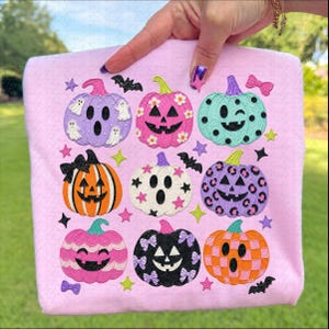May include: A light pink t-shirt featuring a grid of nine colorful pumpkins with various patterns and expressions. The design includes ghosts, stars, bats, and bows in shades of purple, orange, black, white, pink, and teal, arranged in a square.