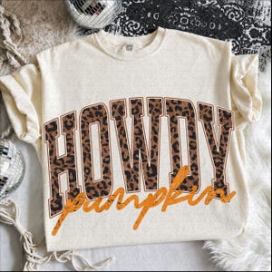 Puede incluir: Una camiseta de manga corta color crema con la palabra HOWDY en letras grandes de estilo bloque con un estampado de leopardo marr&oacute;n y negro. Debajo de la palabra HOWDY, la palabra pumpkin est&aacute; escrita en cursiva naranja desgastada.