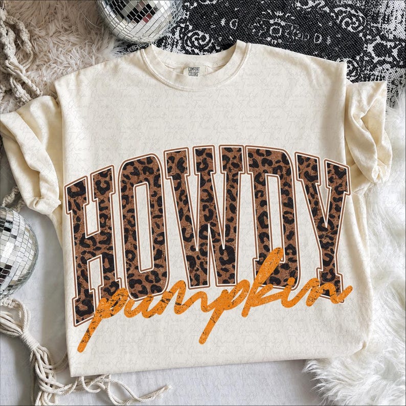 Puede incluir: Una camiseta de manga corta color crema con la palabra HOWDY en letras grandes de estilo bloque con un estampado de leopardo marr&oacute;n y negro. Debajo de la palabra HOWDY, la palabra pumpkin est&aacute; escrita en cursiva naranja desgastada.