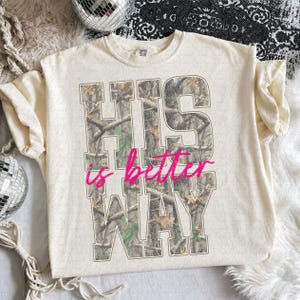 K&ouml;nnte beinhalten: Ein cremefarbenes Kurzarm-T-Shirt mit gro&szlig;en Blockbuchstaben, die HIS WAY mit einem Wald-Tarnmuster-F&uuml;llung zeigen. Die W&ouml;rter is better sind in einer leuchtend pinkfarbenen Schreibschrift &uuml;ber die Mitte des Designs gedruckt.