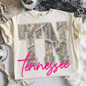 May include: A cream-colored short-sleeve t-shirt featuring the large block letters TN filled with a camouflage forest pattern. Overlaid across the bottom of the letters is the word Tennessee written in a bright pink cursive script font.