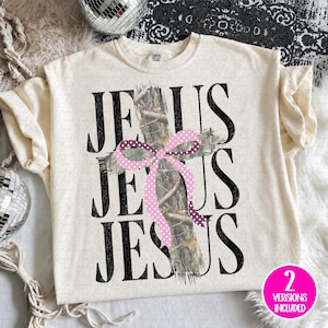 Könnte beinhalten: Ein cremefarbenes Kurzarm-T-Shirt mit dem dreimal in großen schwarzen Buchstaben aufgedruckten Wort JESUS. Ein Kreuz mit Tarnmuster ist hinter dem Text zentriert und mit einer rosa-weiß gepunkteten Schleife verziert.