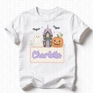 May include: A white short-sleeve t-shirt featuring a Halloween-themed graphic. The design includes a white ghost, a purple and black haunted house, a carved orange pumpkin, and two black bats. Below, a scalloped label displays the name Charlotte.
