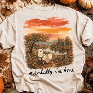 May include: A cream-colored t-shirt featuring a graphic of a ghost sitting in a wooden chair by a lake, holding a cup. The scene includes autumn trees, a house, and a sunset sky. The text "mentally i'm here" is printed in black below the graphic.