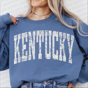 May include: A blue sweatshirt with the word "KENTUCKY" printed in distressed white lettering. The sweatshirt is a solid blue color and has long sleeves. The text is in a bold, collegiate-style font.