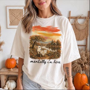 May include: An off-white short-sleeve t-shirt featuring a graphic print of a ghost sitting in a wooden chair by a lake during autumn. The scene includes trees with orange foliage and a small house. The text "mentally i'm here" is printed in black.