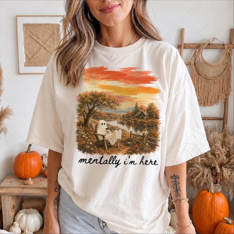 May include: An off-white short-sleeve t-shirt featuring a graphic print of a ghost sitting in a wooden chair by a lake during autumn. The scene includes trees with orange foliage and a small house. The text "mentally i'm here" is printed in black.