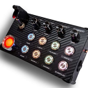 Può includere: Un pannello di controllo nero con una trama in fibra di carbonio, con più pulsanti e manopole. Il pannello include pulsanti illuminati con le etichette "ABS", "MPD", "LIGHT", "FLASH", "PIT-L", "CAM", "RAIN-L", "WIPER" e "S-REPLAY". Sono visibili anche un grande pulsante rosso e un interruttore a levetta.