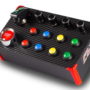 Könnte beinhalten: Ein schwarzer Gaming-Controller mit roten, blauen, gelben und grünen Knöpfen, vier schwarzen Drehknöpfen und einem roten Knopf mit der Aufschrift "Motor Start". Der Controller hat ein Karbonfaser-Muster und rote Einfassungen.