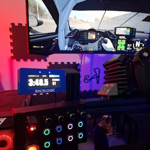 May include: A racing simulator setup featuring a large monitor displaying a car's interior and a racing game. A blue Racelogic display shows lap times. A black control panel with illuminated buttons and a red start button is also visible.