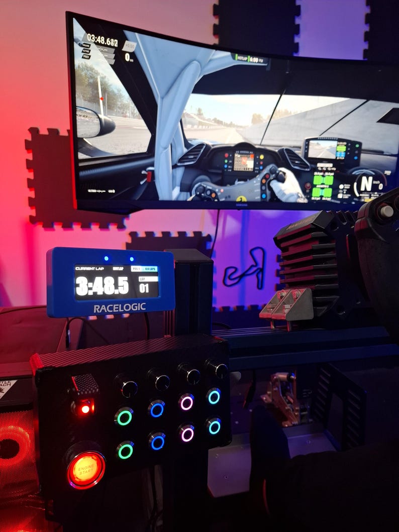 May include: A racing simulator setup featuring a large monitor displaying a car's interior and a racing game. A blue Racelogic display shows lap times. A black control panel with illuminated buttons and a red start button is also visible.