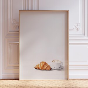 Può includere: Dipinto ad acquerello di un croissant dorato e una tazza di caffè con un piattino bianco. L'opera d'arte è ambientata su uno sfondo neutro, creando un'estetica minimalista. Il dipinto è incorniciato in una cornice di legno marrone chiaro.